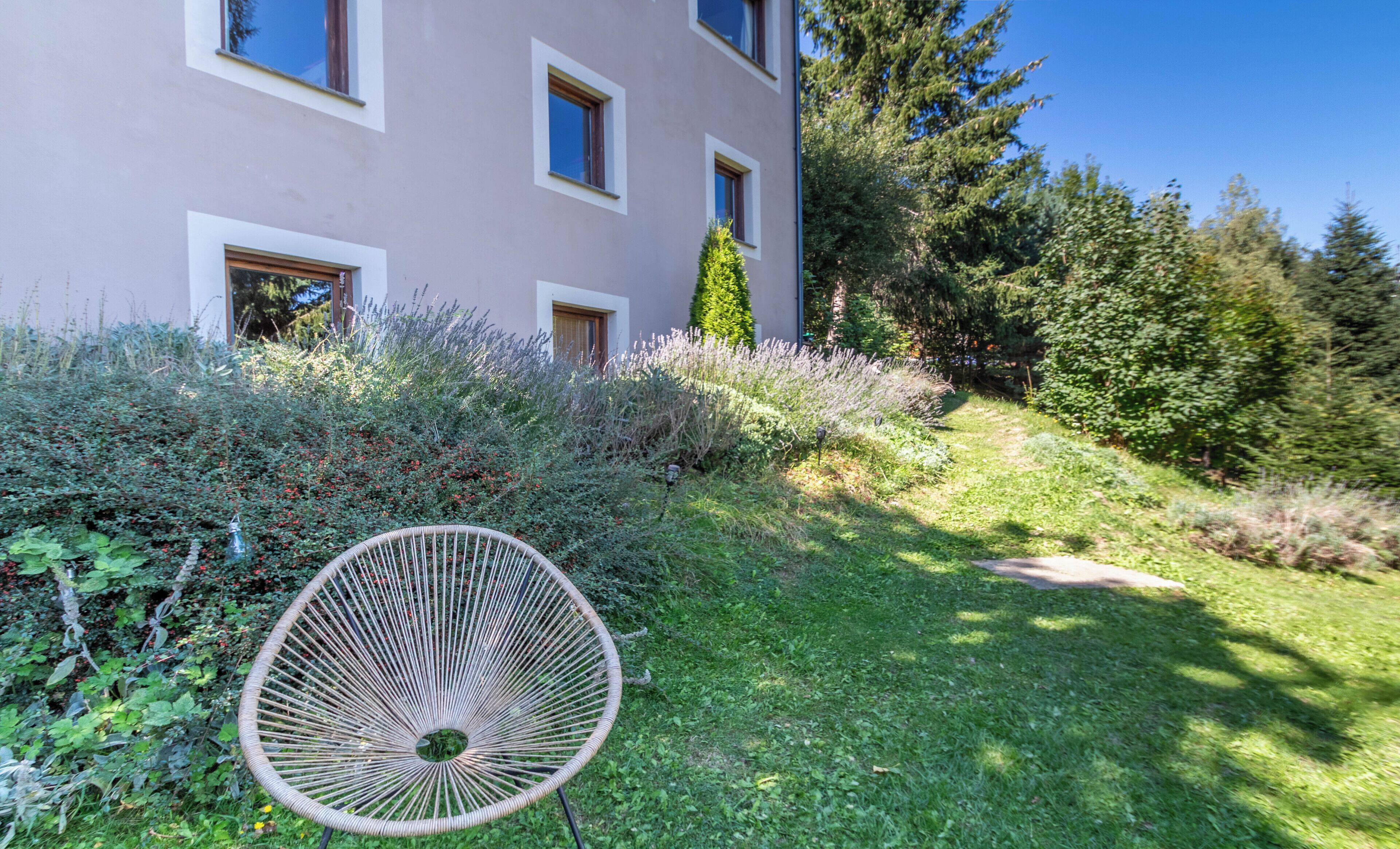 Un edificio moderno con techo plano, amplios ventanales y un jardín cuidado con una silla de mimbre.