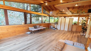 A wooden cabin interior with a wooden bench, exercise bike, and large windows offering a view of trees.