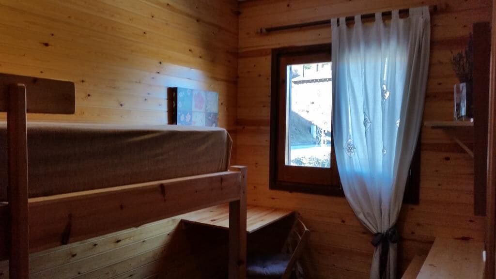 A wooden cabin room with a bunk bed, a window with curtains, and a small shelf with items.