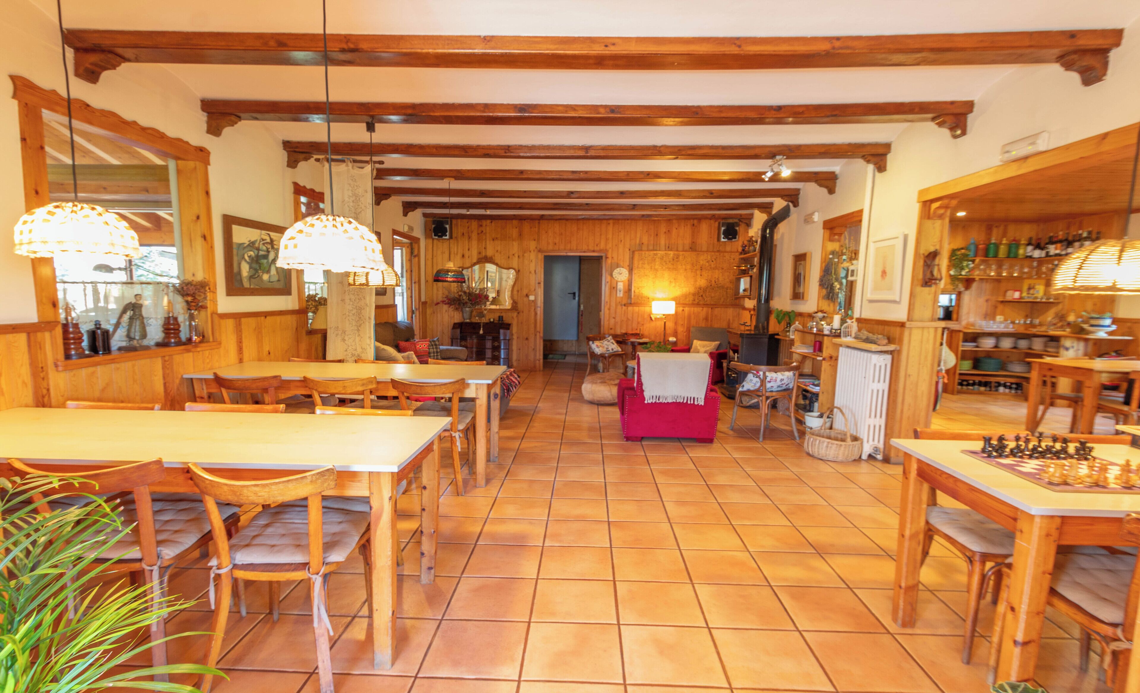 Un comedor amplio con mesas y sillas de madera, un área de estar central con un sofá rojo, y una cocina visible al fondo.