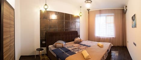 A bedroom with a large bed, a headboard with a textured design, two pendant lights, a small bedside table, and a window with curtains.
