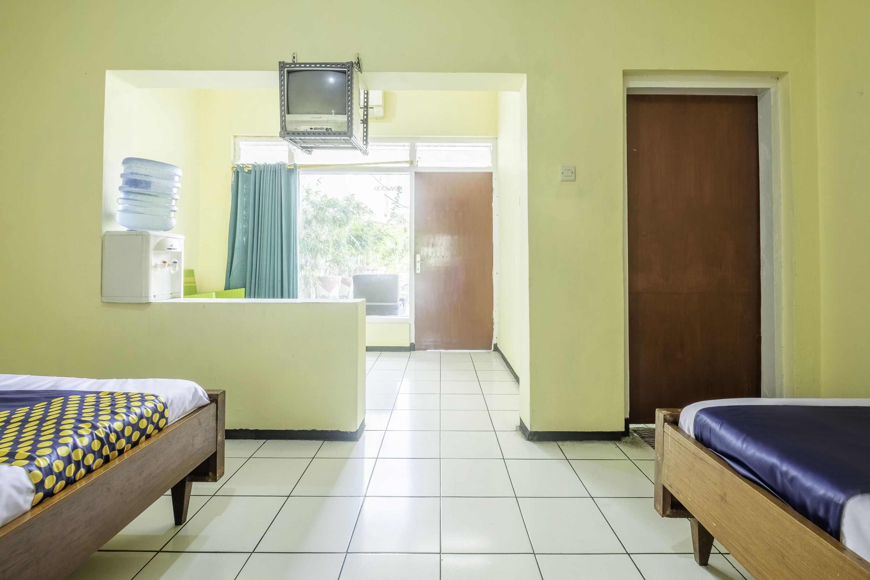 Ein Zimmer mit zwei Betten, einer kleinen Küchenzeile, einem Fenster mit Vorhängen und einer Tür.
