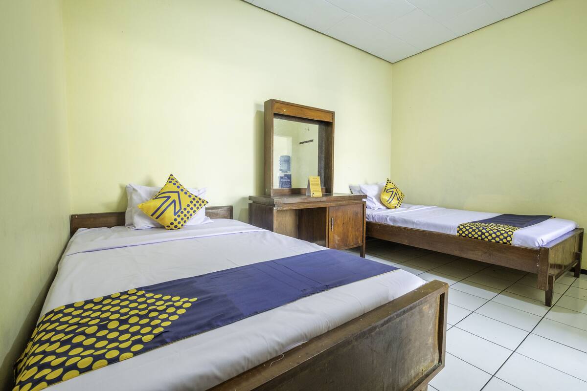 Dos camas individuales con cabeceros de madera, ropa de cama blanca y almohadas estampadas en azul y amarillo. Un escritorio de madera con un espejo y una pequeña estantería se encuentra entre las camas.