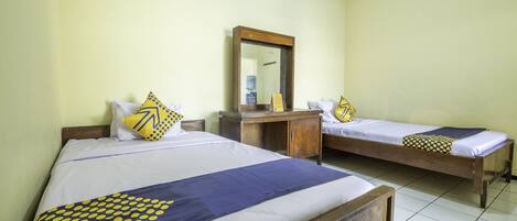 A room with two single beds, each with a wooden frame and white bedding. There is a wooden desk with a mirror above it. The walls are painted a light yellow, and the floor is tiled.