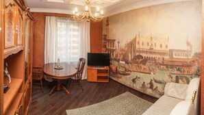 A room with a dining table, chairs, a TV stand, and a large mural of a historical city scene. - Apart Kiev Zlatoustovskaya 50-2 (Kyiv)