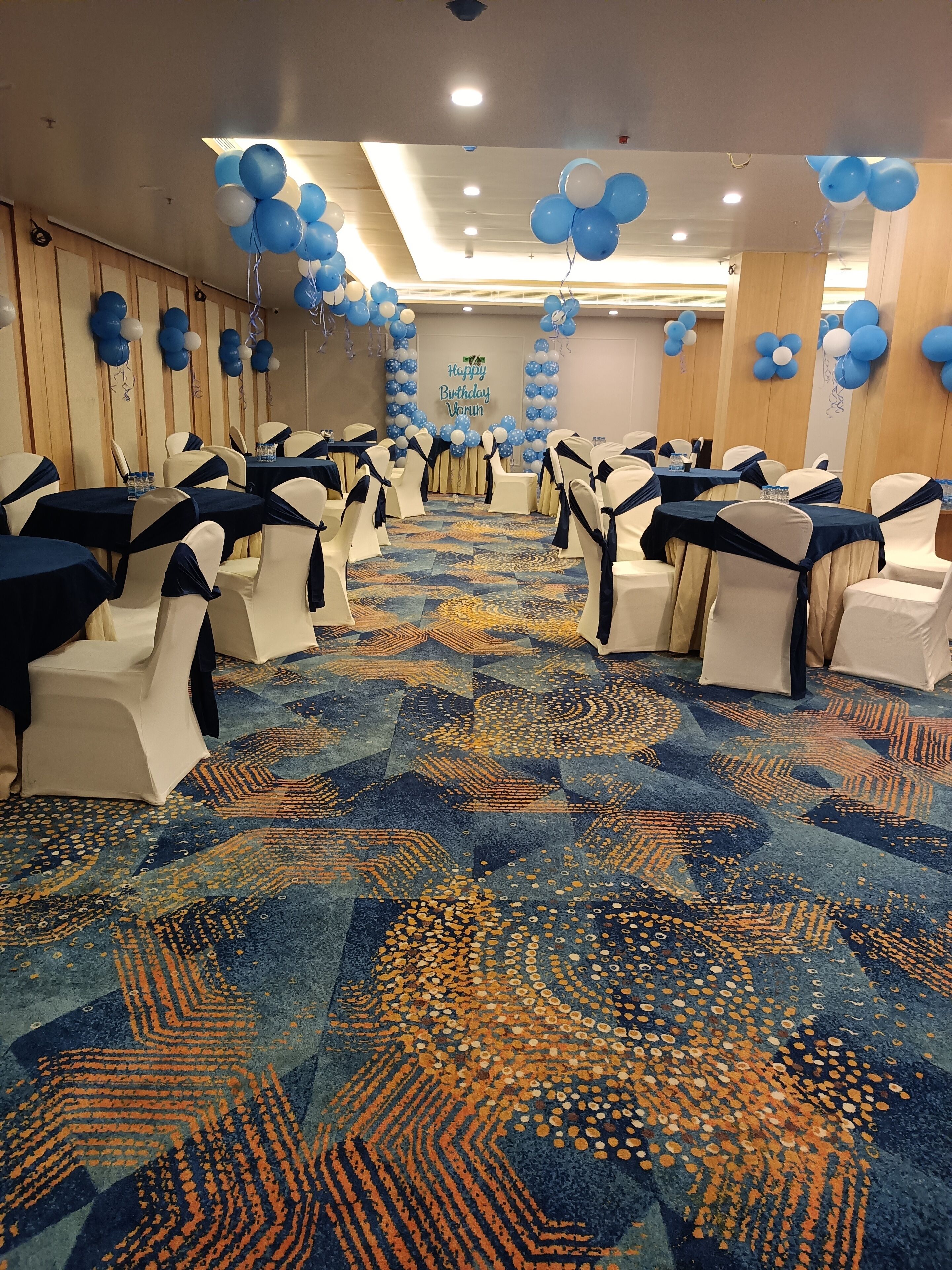 A banquet hall with rows of tables and chairs, blue and white balloons, and a patterned carpet.