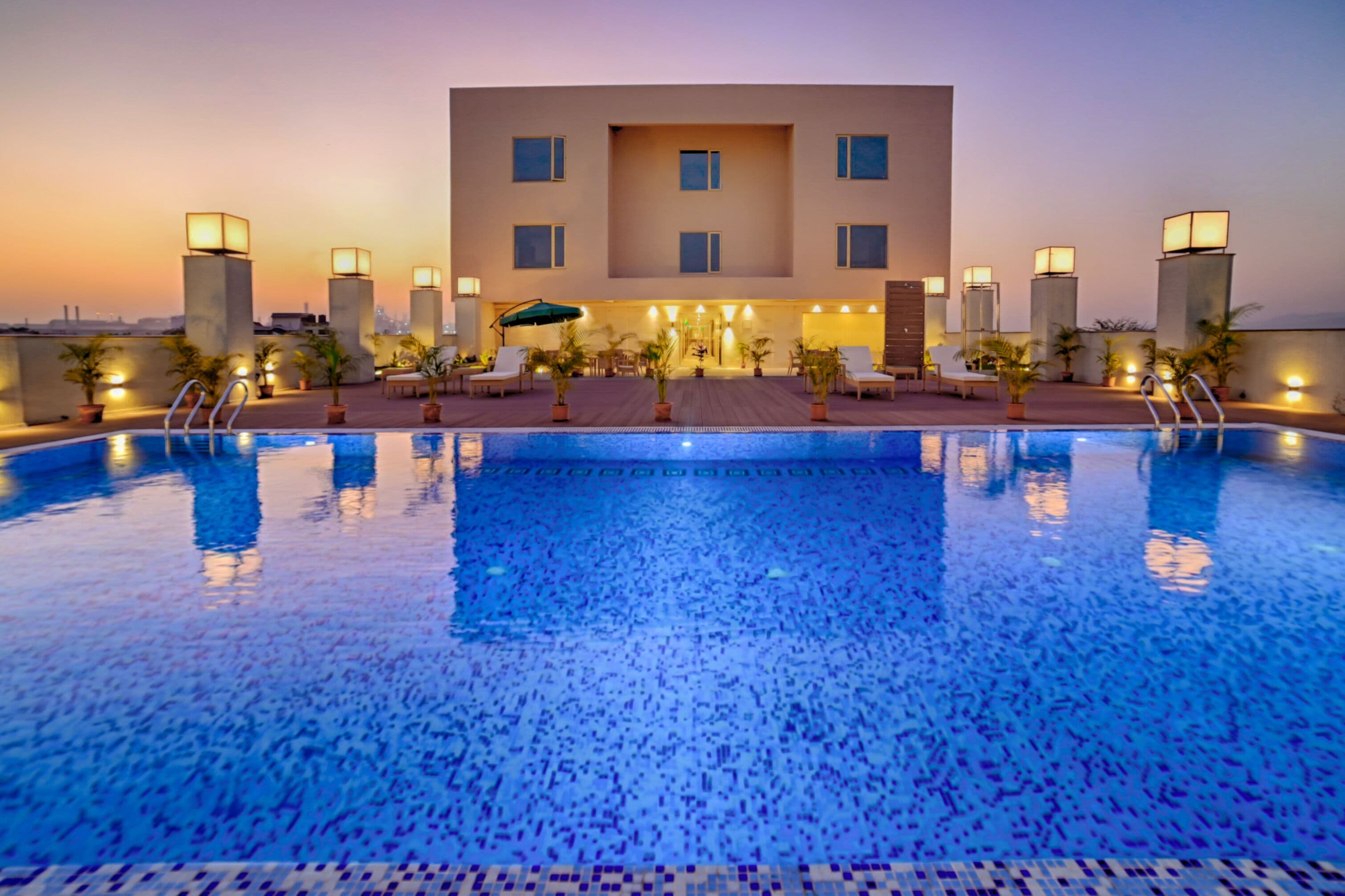 A rooftop swimming pool with a modern building in the background, surrounded by potted plants and outdoor seating.