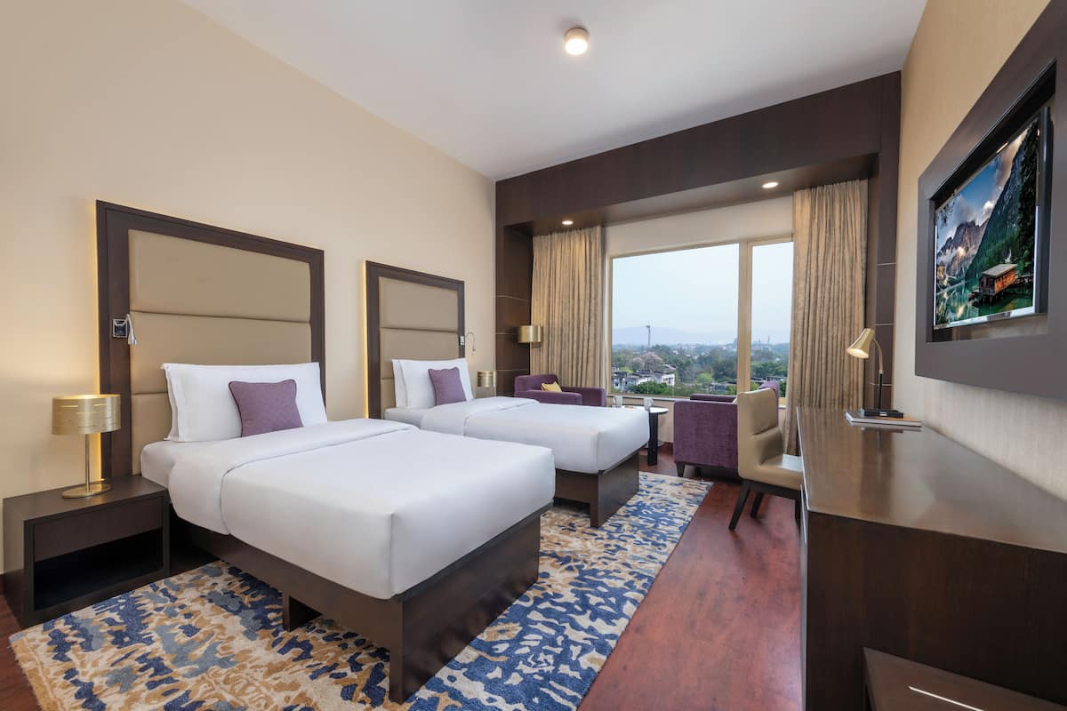 Habitación de hotel con dos camas, un escritorio, una silla, un ventanal con vistas y un televisor de pared.