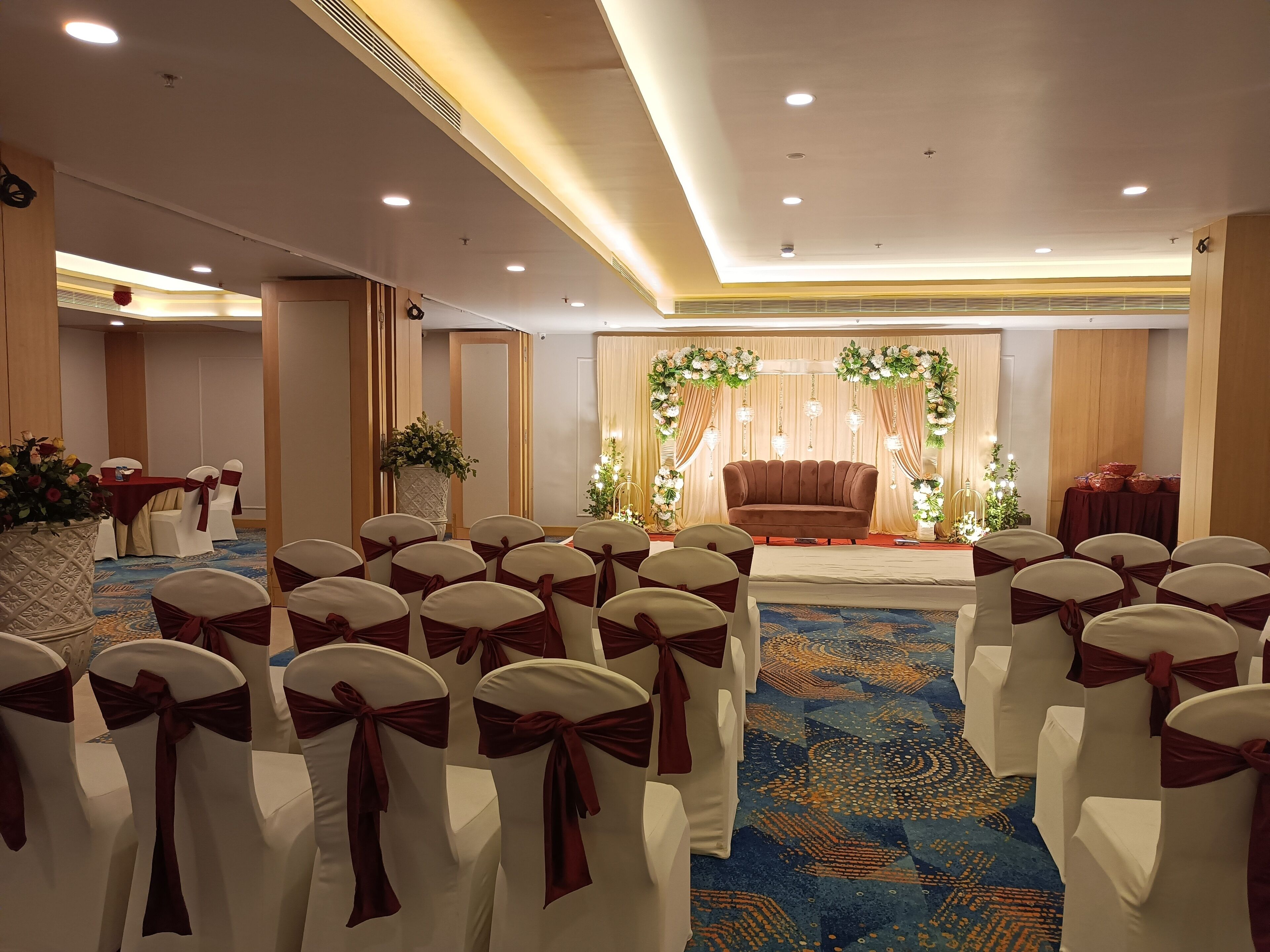 A banquet hall with rows of white chairs with red sashes, a stage with a brown sofa and floral decorations, and a carpeted floor.