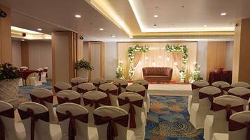 A banquet hall with rows of white chairs with red sashes, a stage with a brown sofa and floral decorations, and a carpeted floor.