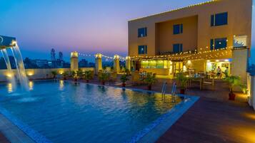 Un área de piscina en la azotea con fuente de agua, mobiliario exterior e iluminación con luces colgantes.