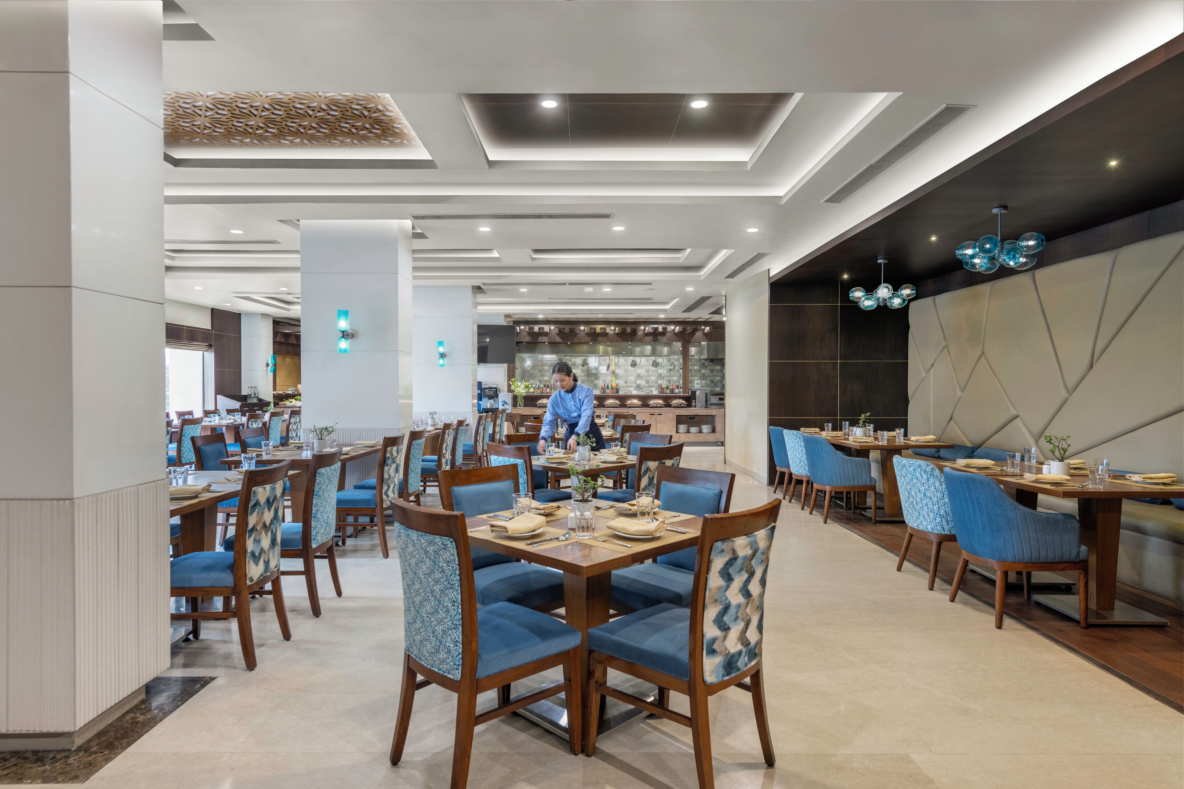 Een modern restaurant met houten tafels en blauwe bekleden stoelen, een bar en een decoratief plafond.