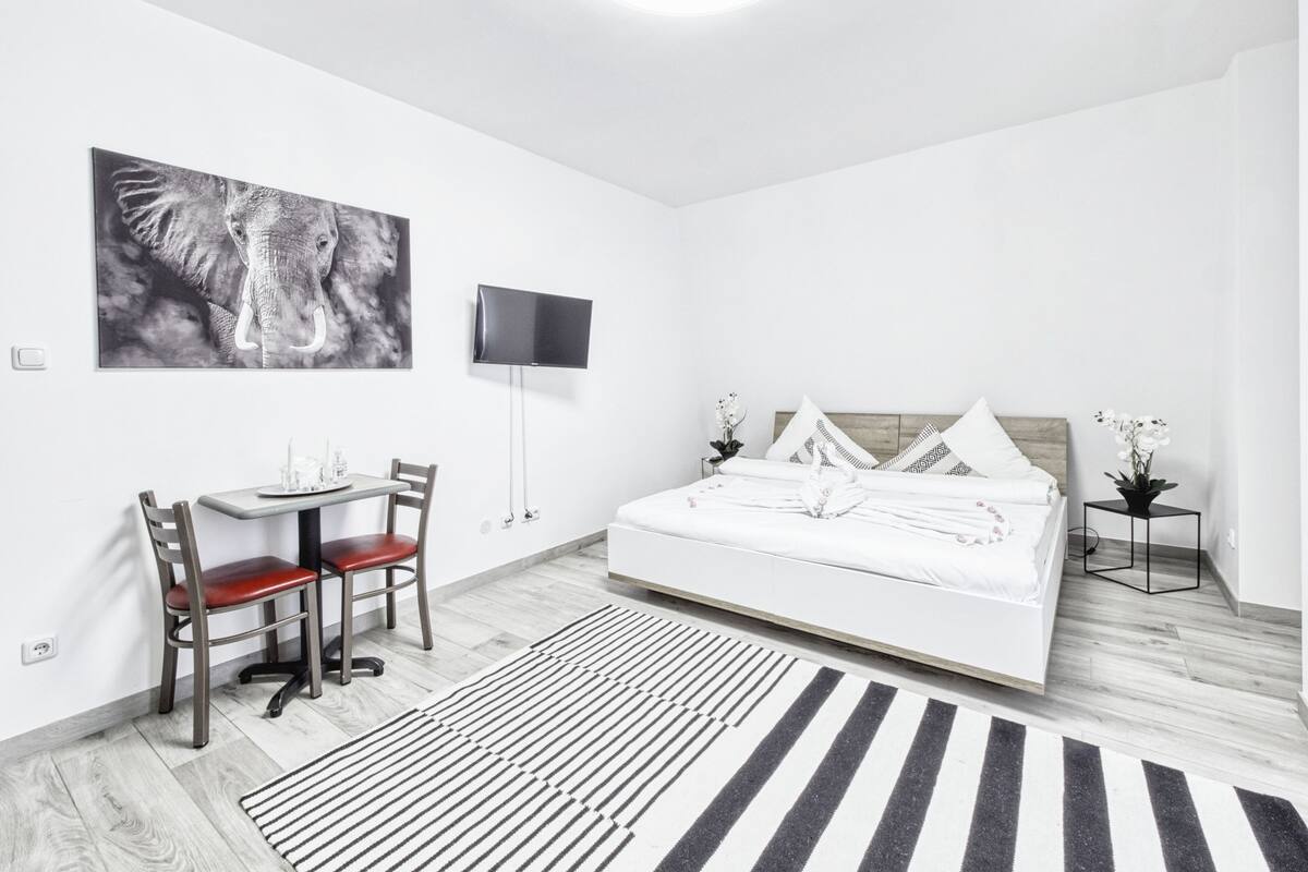 A modern hotel room with a large bed, a desk with a chair, a flat-screen TV mounted on the wall, and a large elephant artwork.