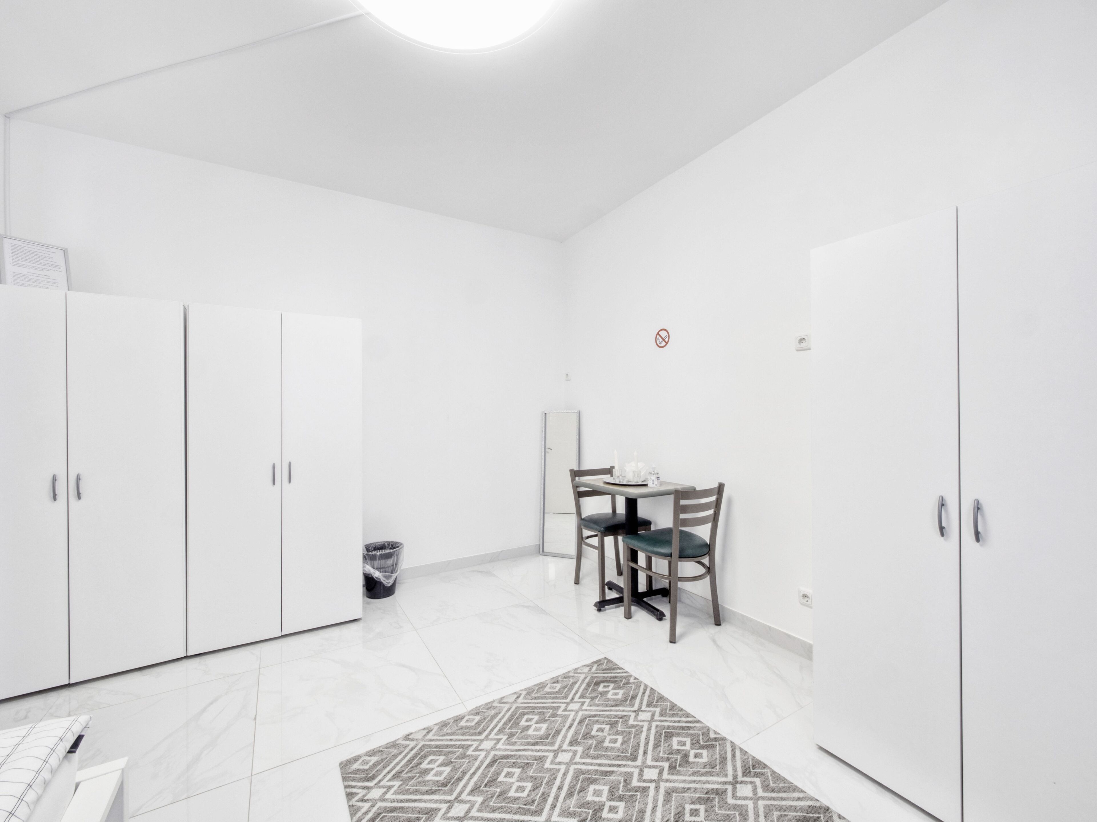 A modern, minimalistic room with white walls and a tiled floor. It includes a dining area with a table and chairs, and a wardrobe with two doors.