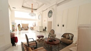 A spacious living room with a chandelier, a round coffee table, and a sofa. There are two armchairs and a patterned ottoman. The room has a large window and a door leading to another area. - Apatel The Mansion Tower Capilano (Jakarta)