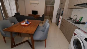A modern kitchen with a built-in washing machine, a dining area with a wooden table and grey chairs, and a small seating area with a TV. - Apatel The Mansion Tower Capilano (Jakarta)