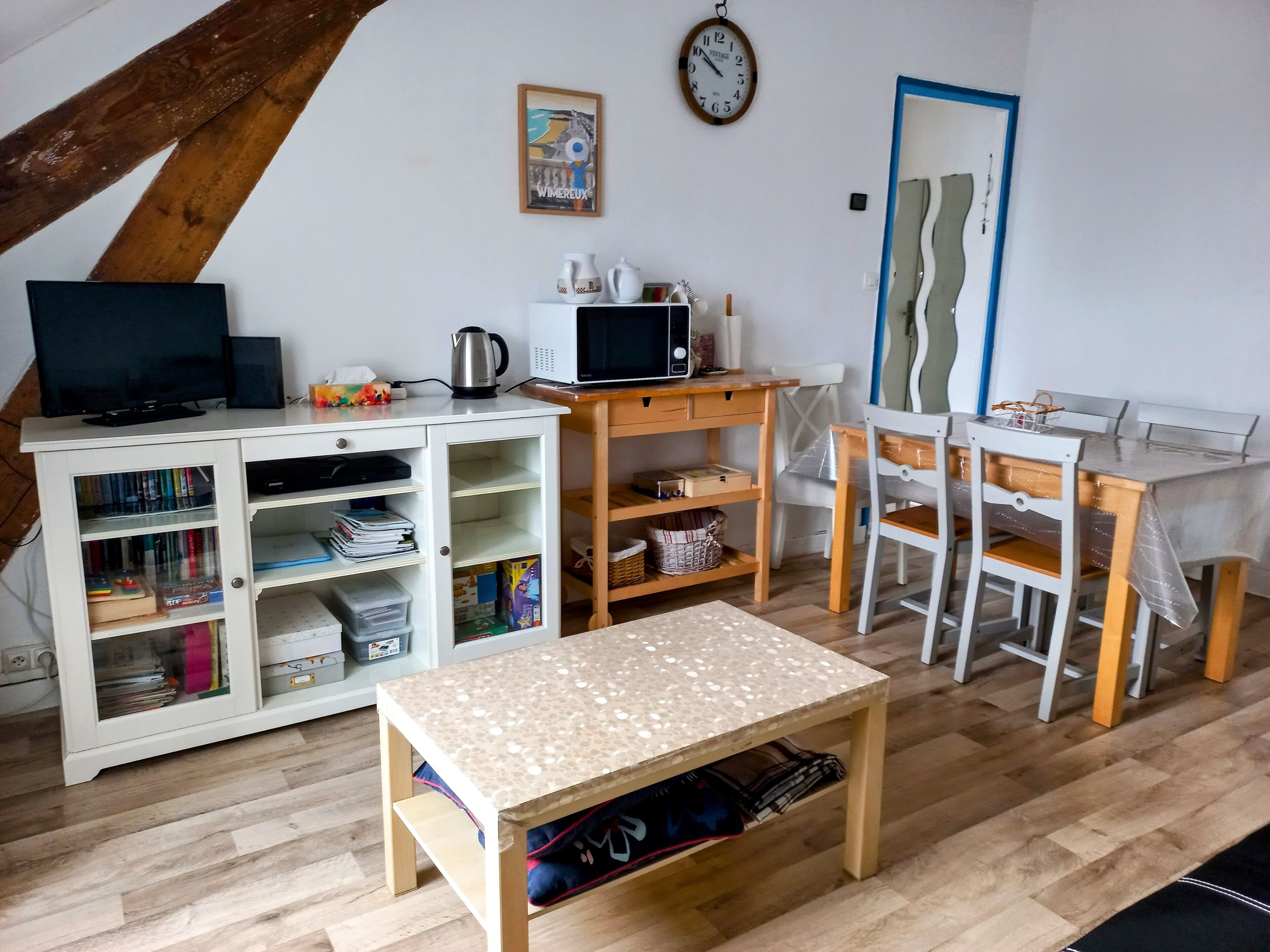 A cozy living room with a TV, a coffee maker, a microwave, a bookshelf, a dining table with chairs, and a wooden floor.