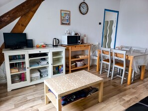 A cozy living room with a TV, a coffee maker, a microwave, a bookshelf, a dining table with chairs, and a wooden floor.