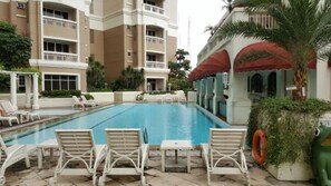 A swimming pool with lounge chairs and a small table, surrounded by a building with balconies and a palm tree. - Apatel Apartement Kedoya Elok (Jakarta)
