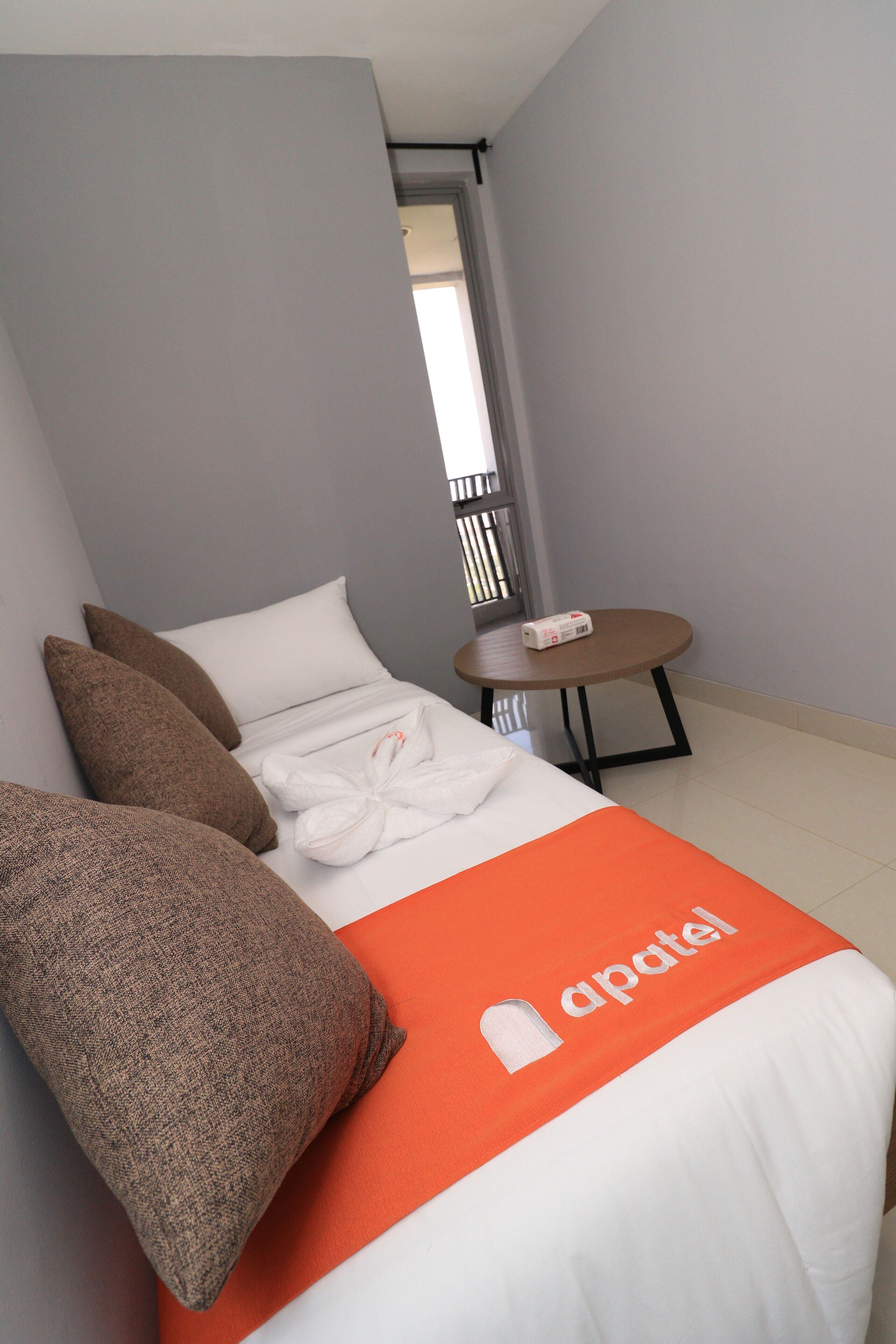 A neatly made bed with a white and orange bedspread, two brown pillows, and a white towel. There is a small round table with a white object on it, and a window with blinds in the background.