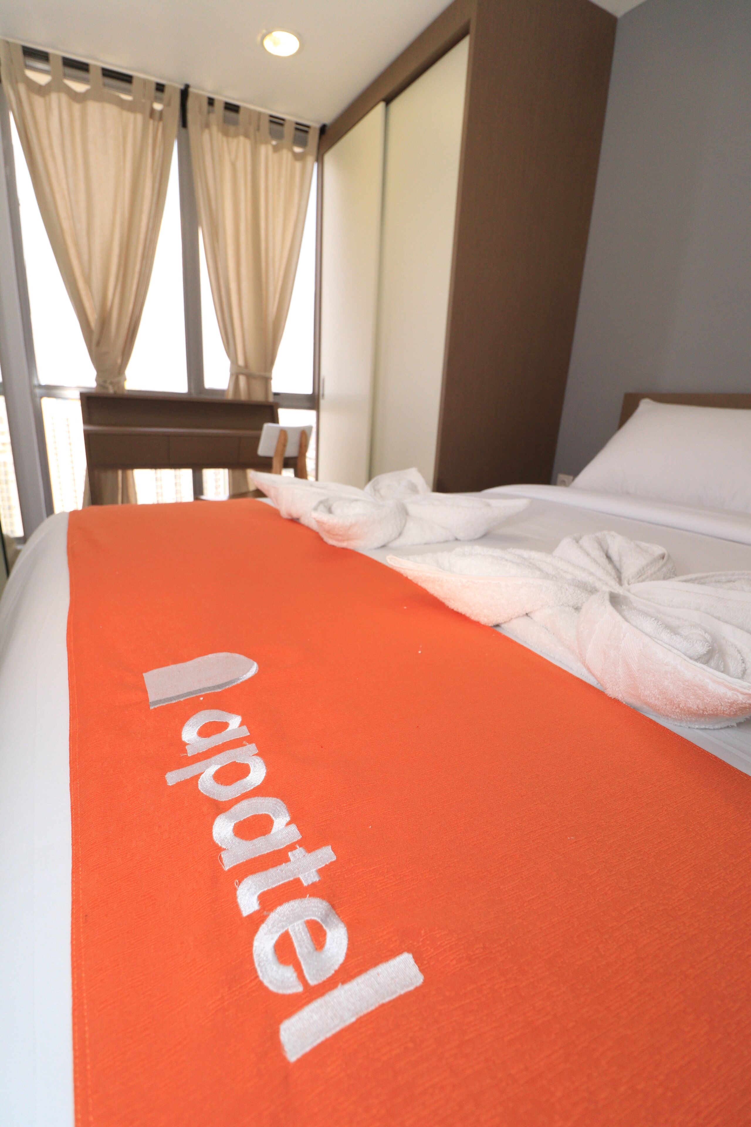 Habitación de hotel con una cama cubierta con una manta naranja y la palabra 'hotel' visible, una ventana con cortinas y un cabecero de madera.