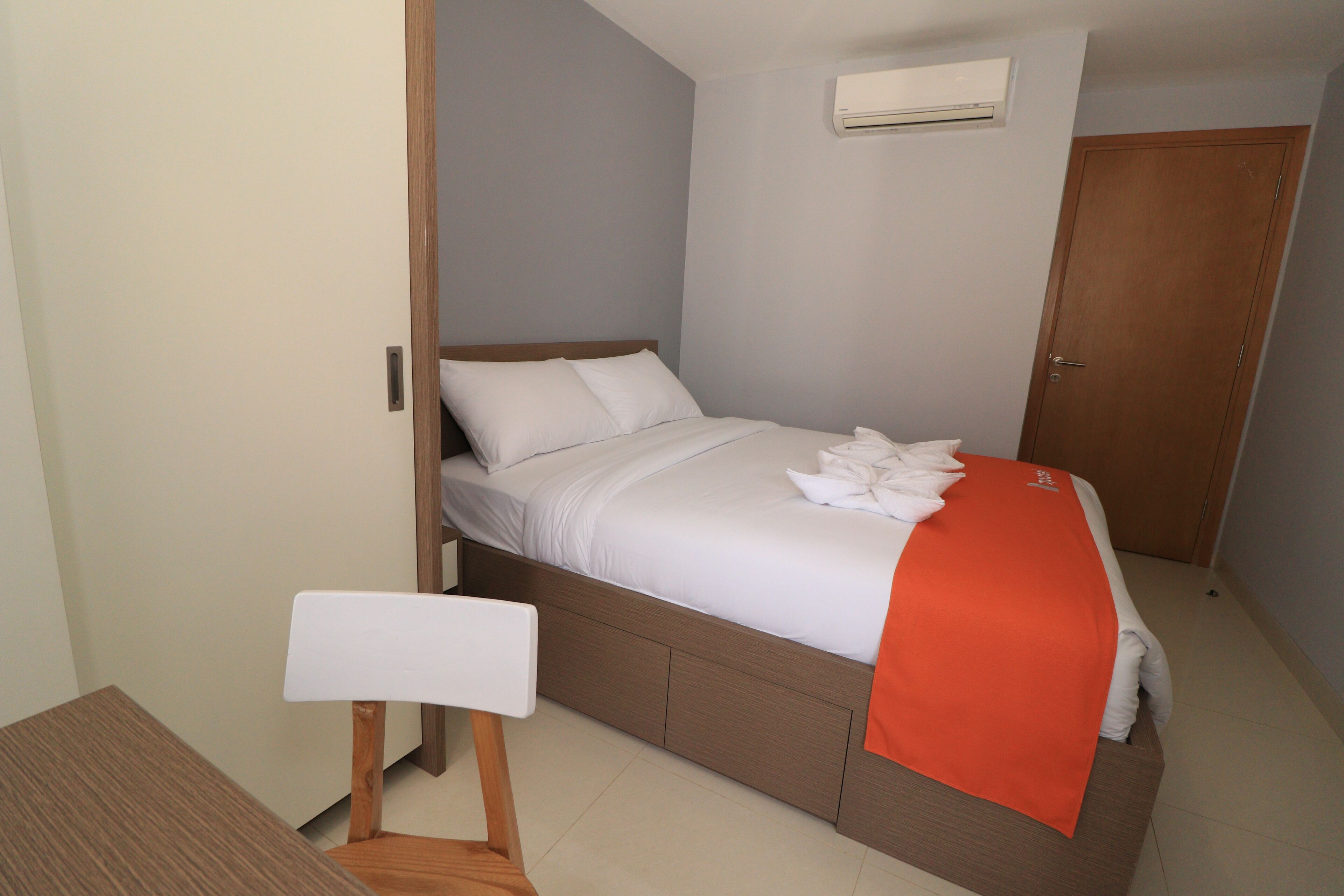 A neatly made bed with a white comforter and pillows, a wooden bedside table, and a small chair.