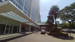 Um edifício moderno com fachada de vidro, entrada coberta e estacionamento com carros e cones de trânsito.