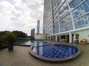 A modern building with a large glass facade, a sign for 'Sogo', and a pool area with lounge chairs. - Apatel U Residence 1 Lt. 10 (Curug)
