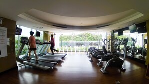 Academia com esteiras, bicicletas ergométricas e uma grande janela com vista para vegetação.