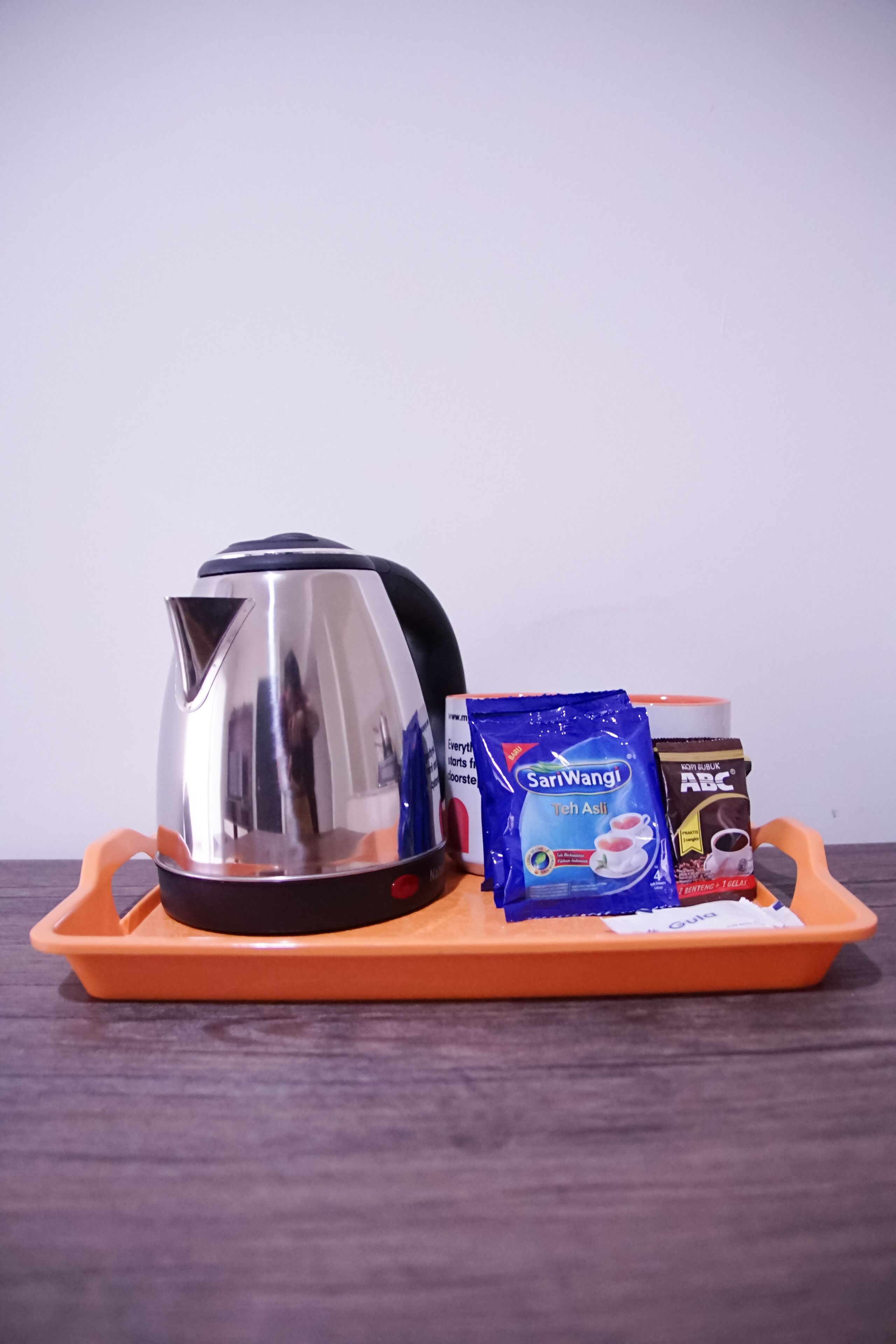 En vattenkokare i rostfritt stål, ett blått paket med SariWangi-te och ett paket med snabbkaffet ABC, placerat på ett orangefärgat bricka.