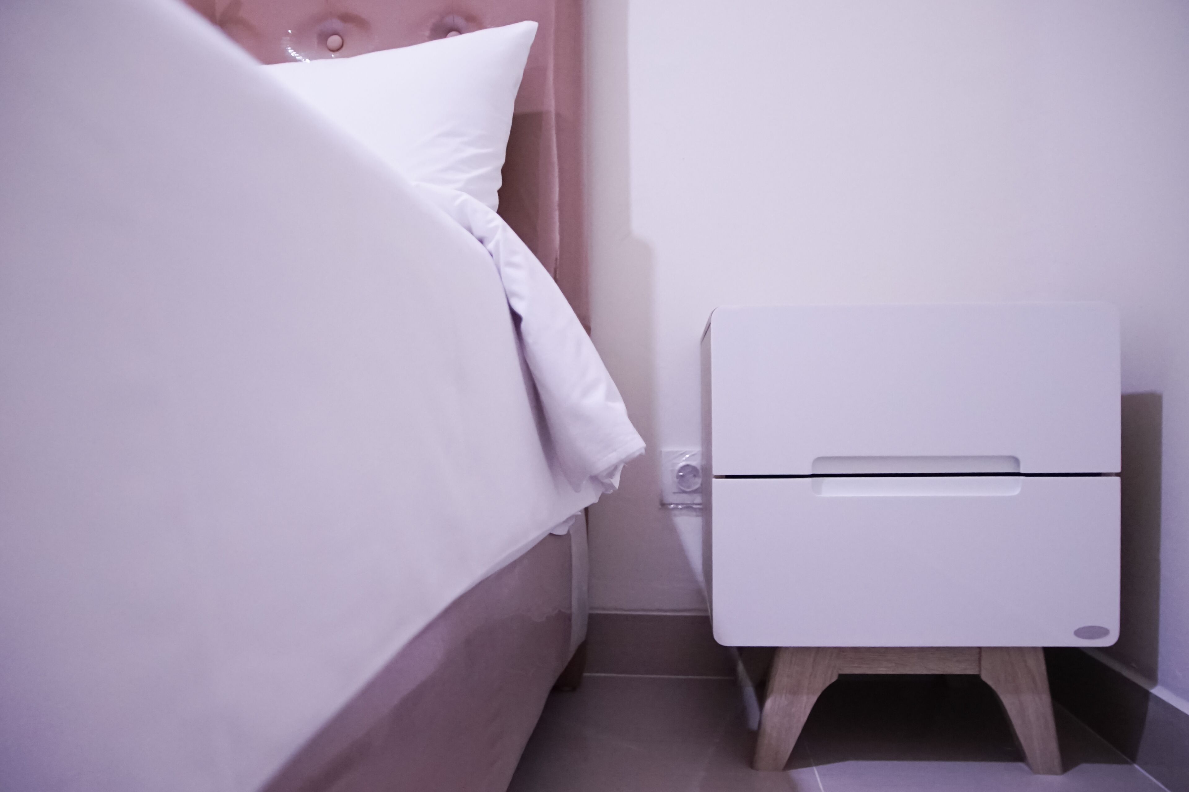A small, neatly made bed with white linens and pillows, a white bedside table with a drawer, and a small wooden stool.