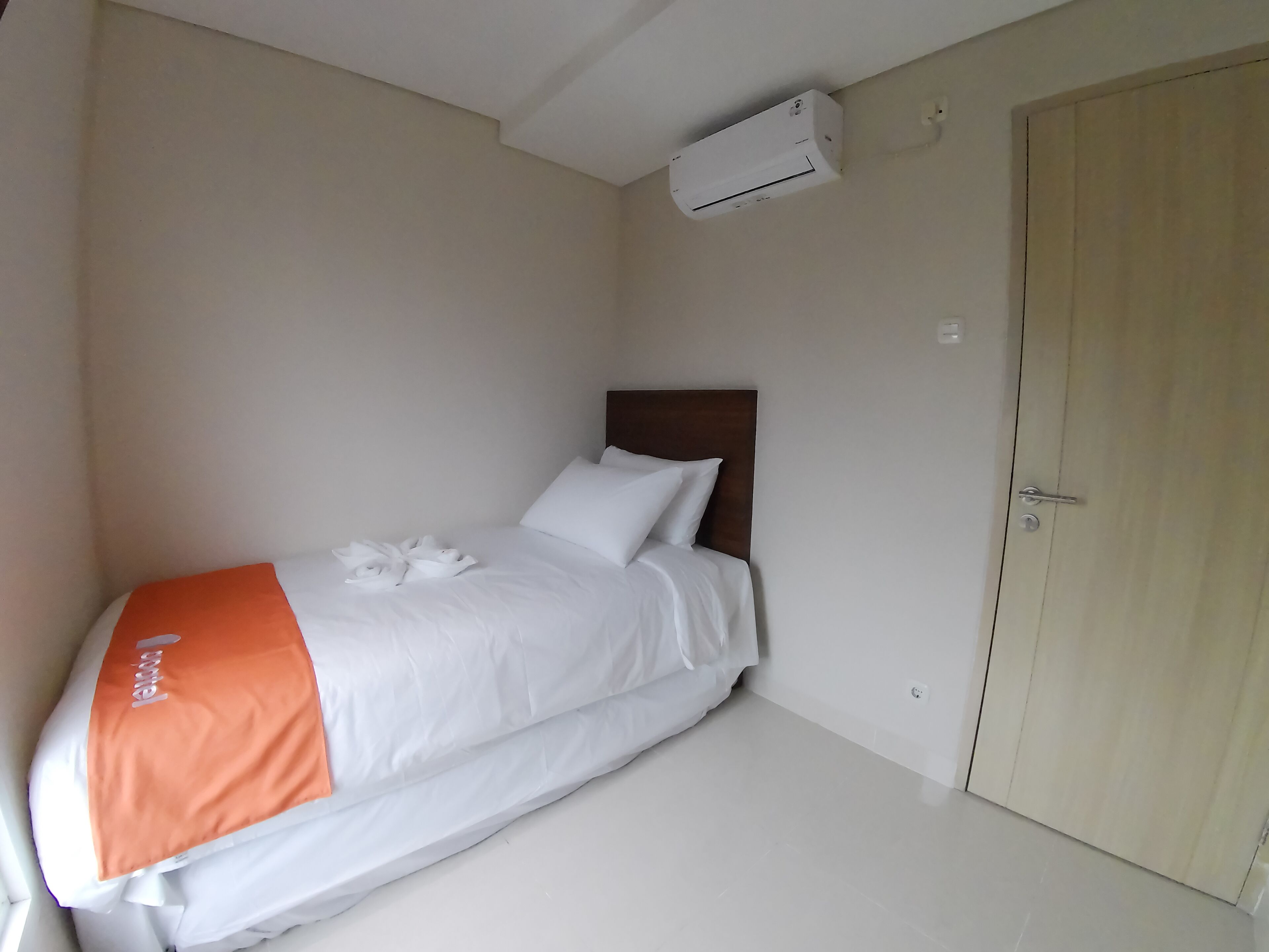 Un lit simple avec une literie blanche et orange, une tête de lit en bois et un appareil de climatisation blanc fixé au mur.