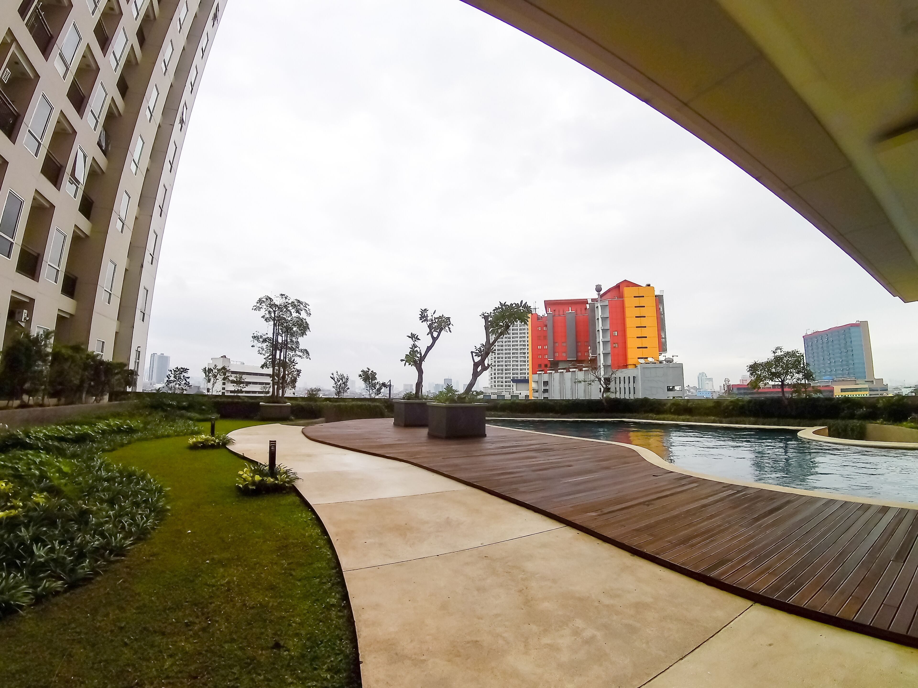 A modern urban landscape with a curved pathway leading to a water feature, surrounded by greenery and under a bridge.