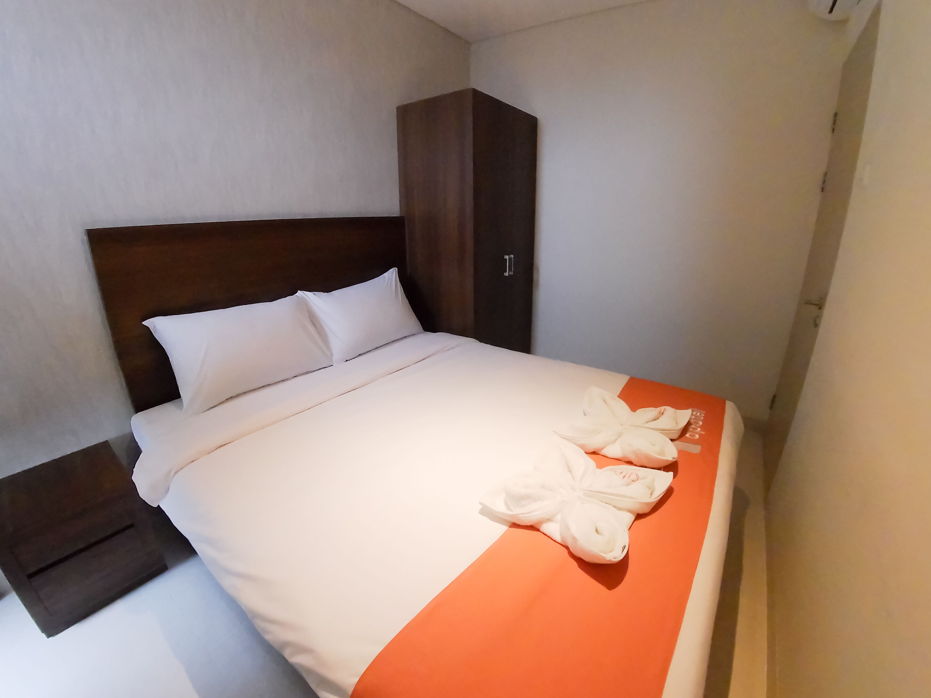 Un lit bien fait, avec du linge de lit blanc et une touche de couleur orange, une tête de lit en bois, une table de chevet et une armoire.