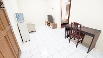 Una habitación pequeña y limpia con suelo de baldosas, un armario de madera, un dispensador de agua, un televisor sobre un mueble de madera, un escritorio con una silla y un espejo en la pared.