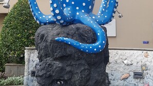 En blå bläckfiskstaty med vita prickar, placerad på en stenbas i en innergård med en byggnad i bakgrunden.