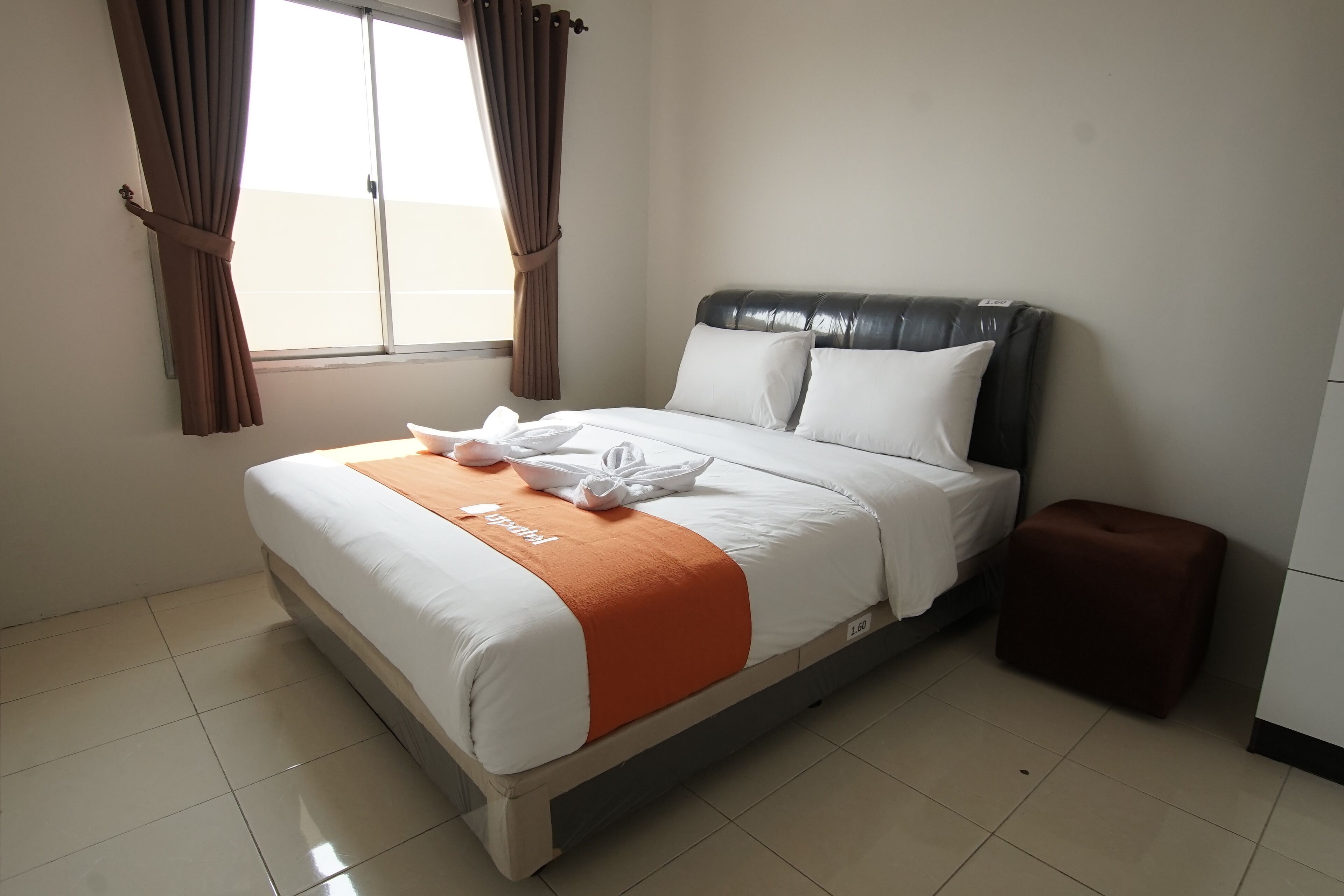 Una cama bien hecha con ropa de cama blanca y naranja, un reposapiés marrón y una ventana con cortinas.