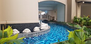 A pool with a mosaic tile design, surrounded by greenery and stone sculptures.