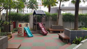 A children's play area with slides and a climbing structure, surrounded by greenery and a checkered floor. - Apatel Silkwood Residences Alam Sutra (Tangerang)