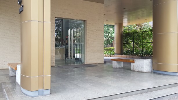 A modern building entrance with a glass door, beige pillars, and a bench. - Apatel The OAK Tower Pulo Gadung (Jakarta)