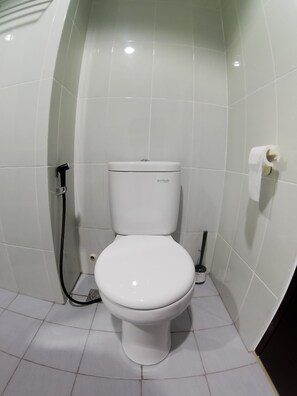 A white toilet with a flush handle, a toilet paper holder, and a bidet attachment in a bathroom with white tiled walls and floor.
