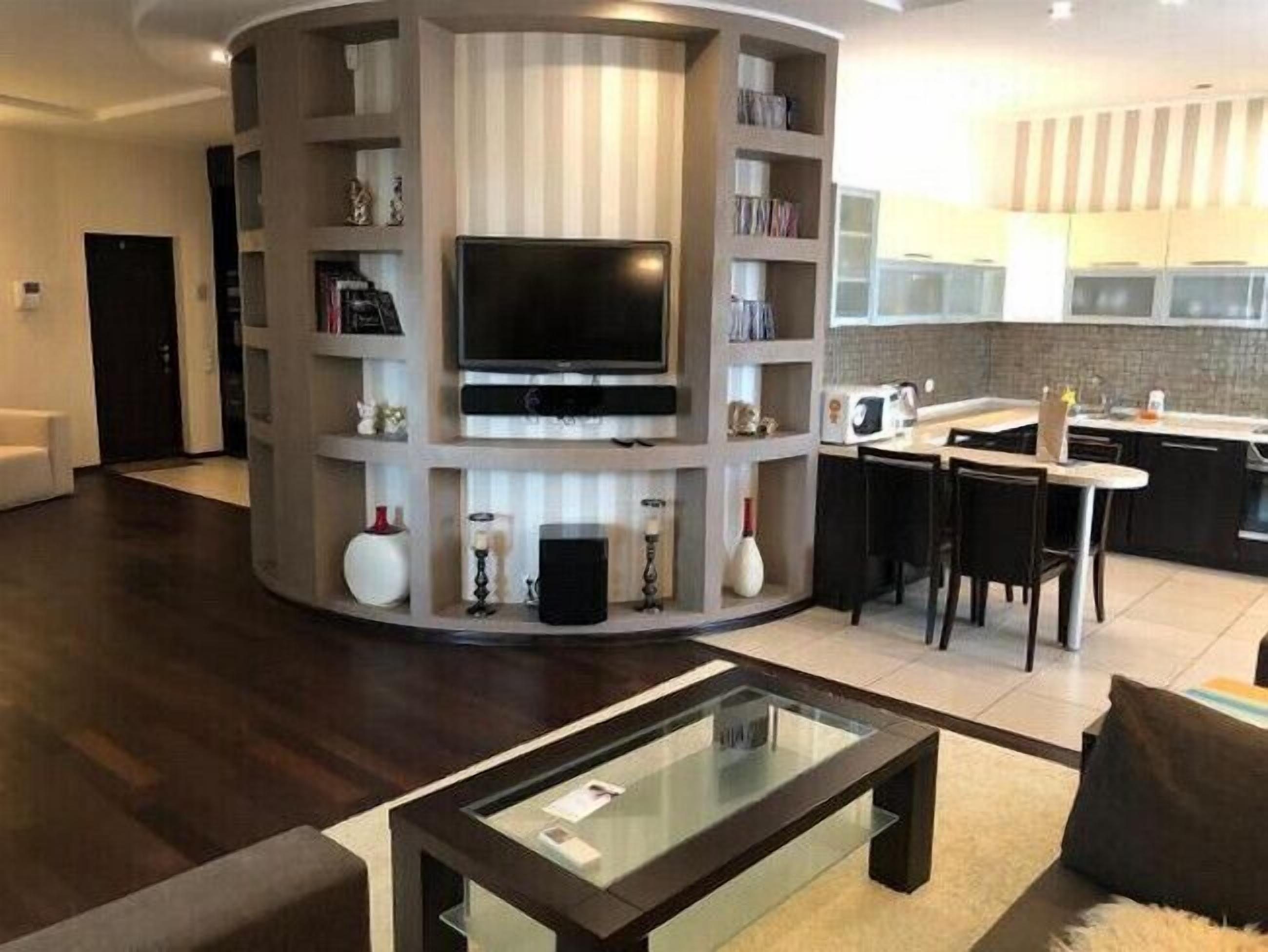A modern living room with a built-in TV, a glass coffee table, and a kitchen area with a microwave and bar stools.
