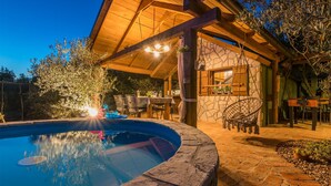 Una piscina de piedra con cubierta de madera, una silla colgante y una mesa con sillas bajo un pérgola de madera.