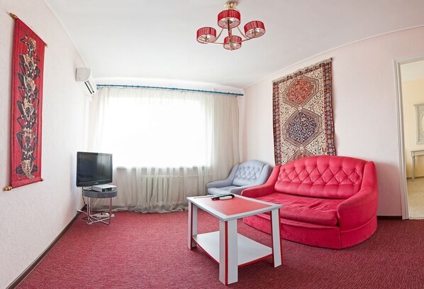 A living room with a red sofa, a white coffee table, a grey armchair, a TV on a stand, and a patterned tapestry on the wall.