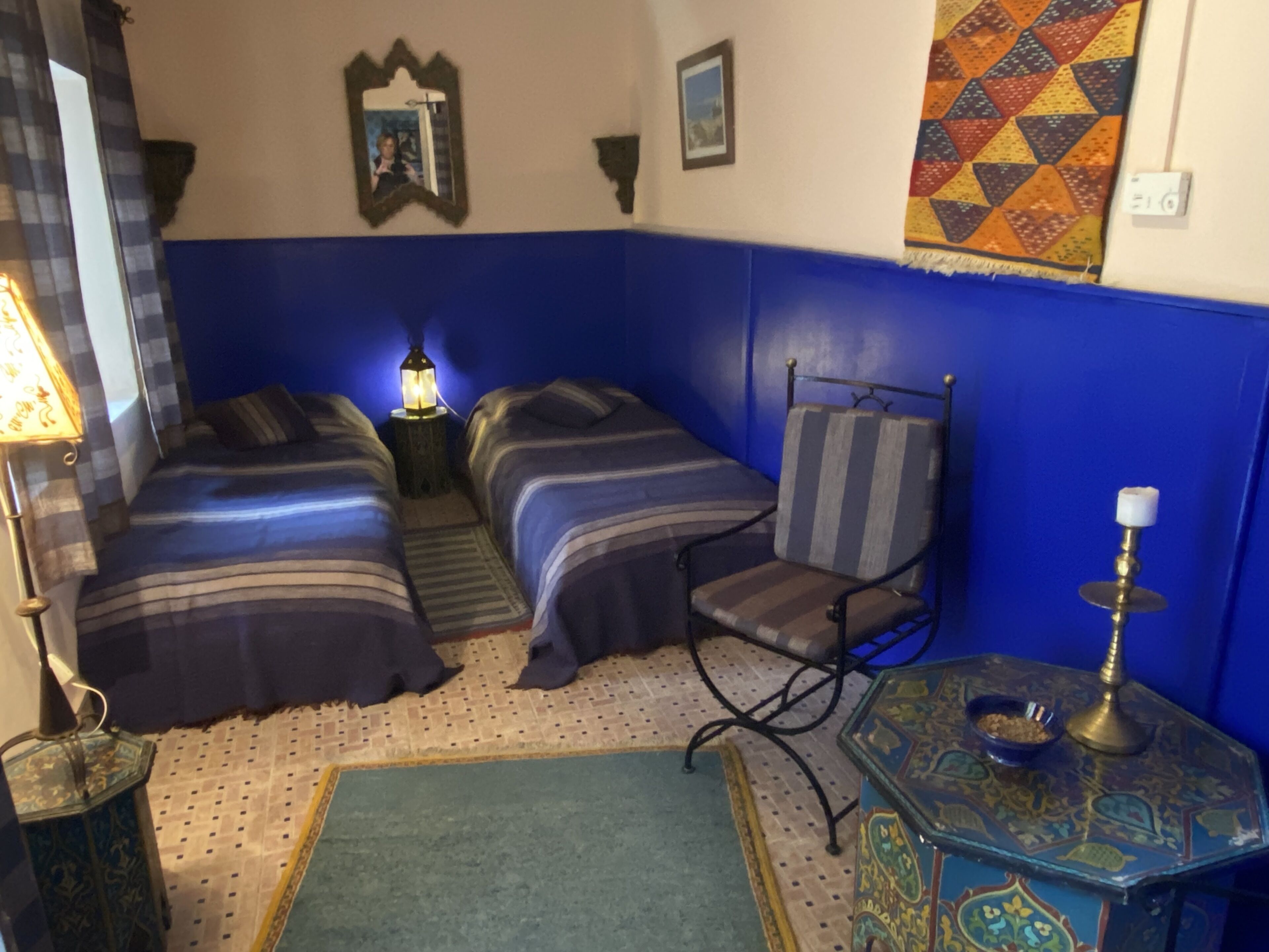 Una habitación con dos camas, una silla, una mesa con lámpara y un tapete estampado.