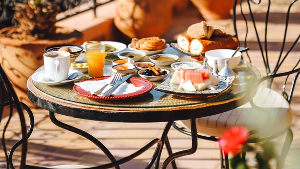 A round table with a variety of breakfast items including pastries, fruits, and beverages, set on an outdoor terrace with potted plants and a metal railing. - Hotel Sherazade (Marrakech)