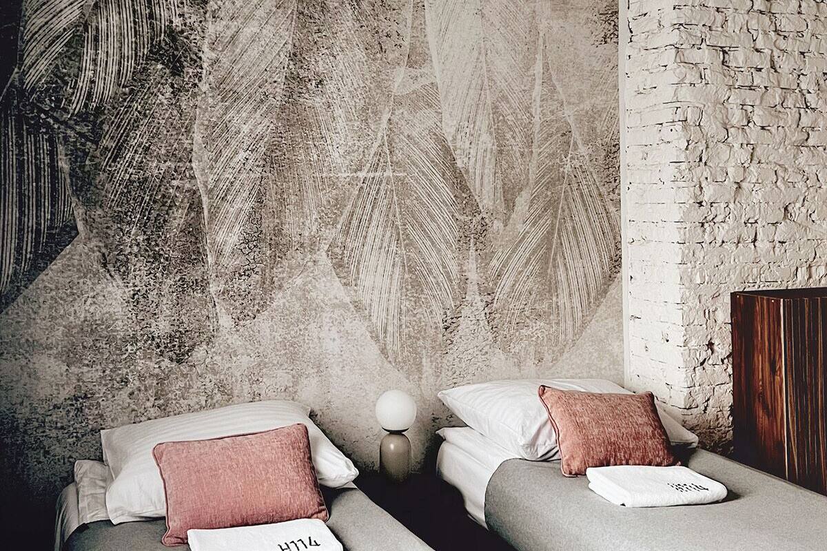 A modern hotel room with two beds, a textured wall, a wooden headboard, and a small bedside table with a lamp.