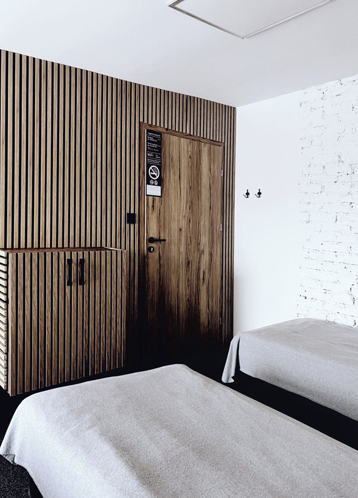 A modern hotel room with a wooden panel wall, a door with a sign, two beds, and a white brick wall.