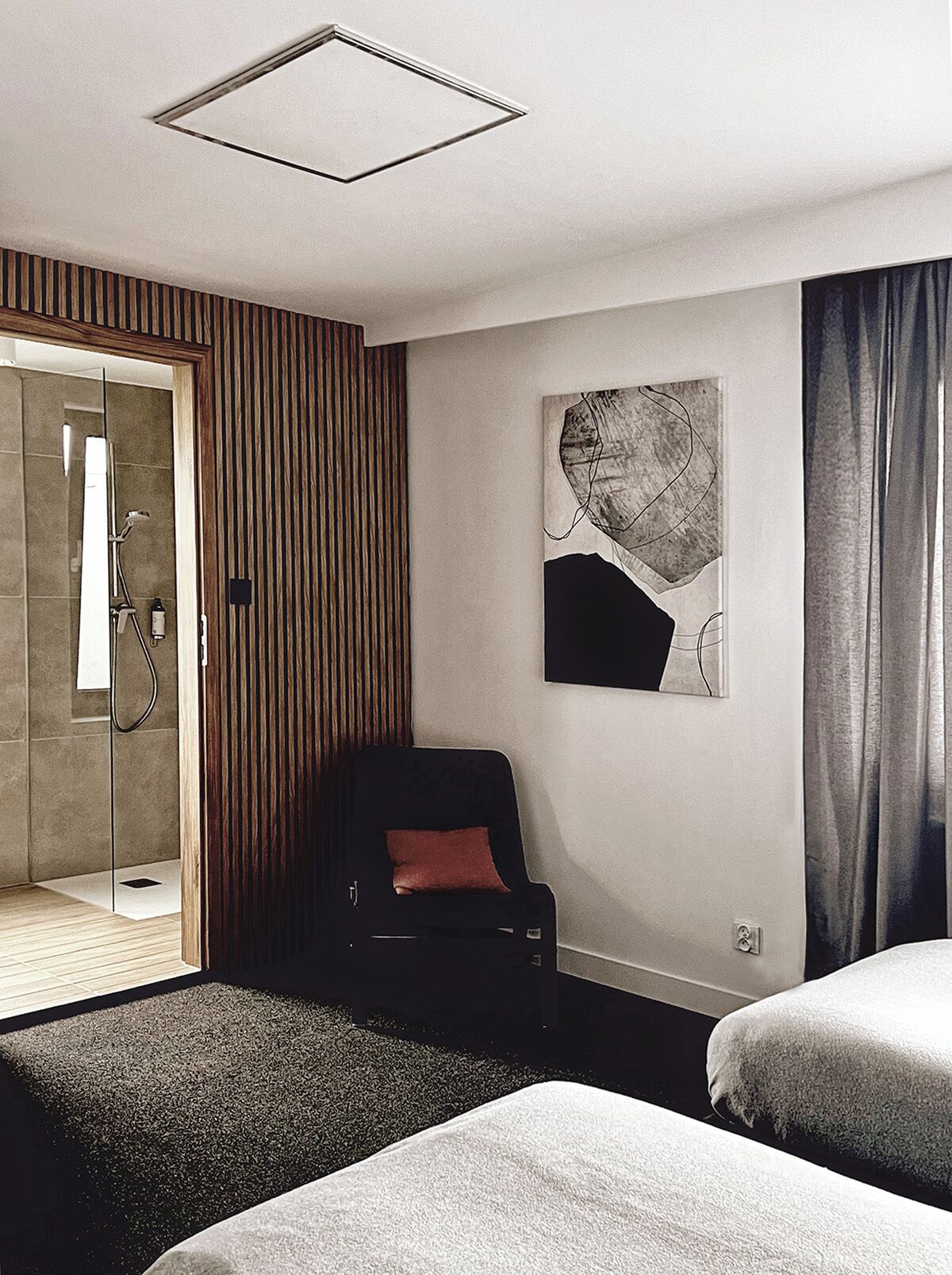A modern hotel room with a glass shower enclosure, a black chair with a red cushion, a large abstract artwork on the wall, and a bed with a grey blanket.