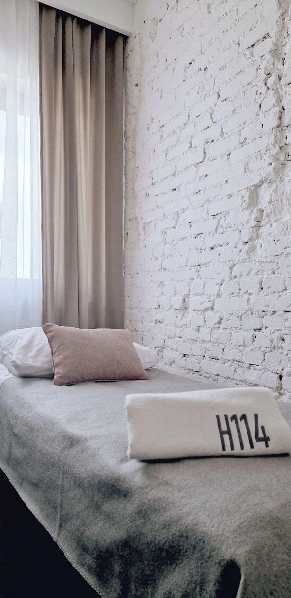 Una habitación con una cama frente a una pared de ladrillo blanco, una ventana con cortinas y una manta con la inscripción 'H114'.